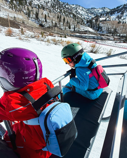 Two people in ski gear sitting on a ski lift in a snowy mountain setting. One person wears a purple helmet and red jacket with a light blue backpack, and the other wears a green helmet and turquoise jacket with a pink backpack.