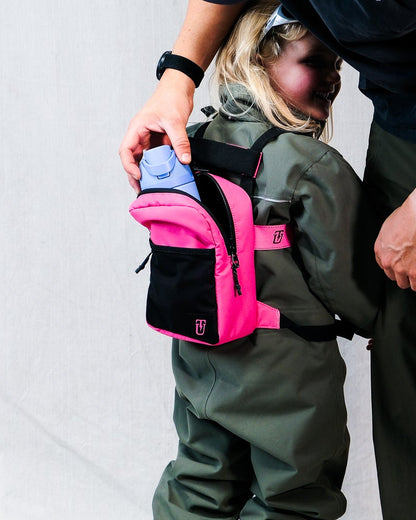A child wearing a bright pink and black ski and snowboard backpack trainer harness, adult putting water bottle inside backpack