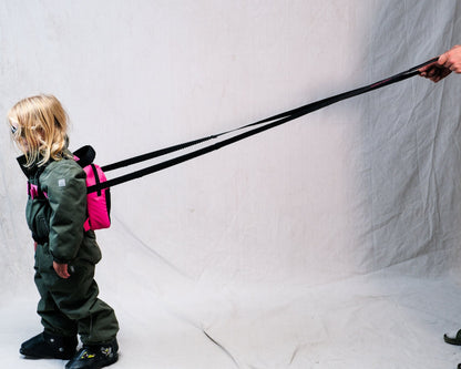 a kid with a pink backpack ski harness is shown from the side while being pulled by an adult holding a black harness attached to the backpack